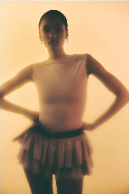 Fashion shot of a woman in a white top and layered skirt posing in front of an amber-colored background, studio setting, medium shot.