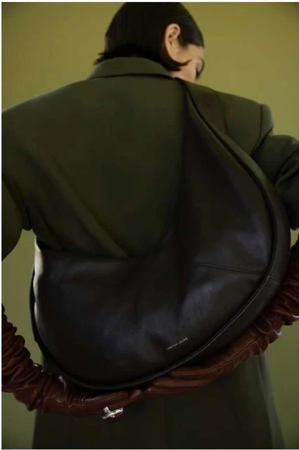 Woman wearing an oversized olive-green jacket and carrying a large, black leather hobo bag with a ruched strap against an olive background.