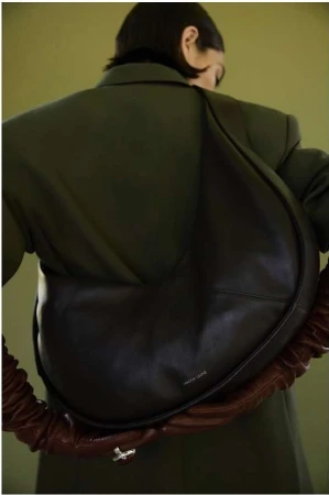 Woman wearing an oversized olive-green jacket and carrying a large, black leather hobo bag with a ruched strap against an olive background.