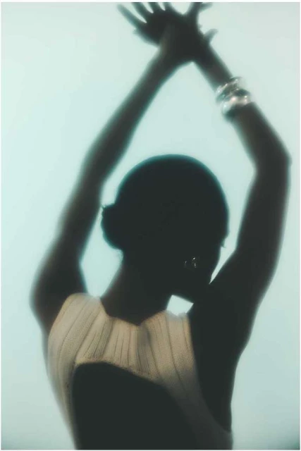 Silhouette of an adult female with arms raised overhead, wearing a light-colored top with an open back, against a blurred background.