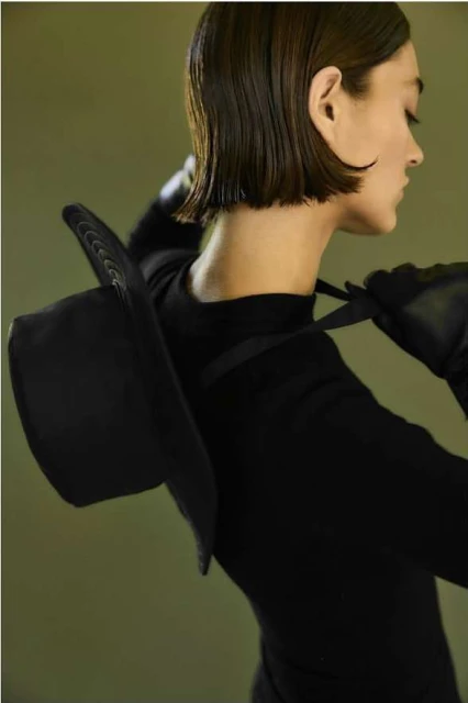 A woman with short hair poses indoors in a black outfit and black gloves, a black hat hanging off her shoulder, profile view against an olive background.