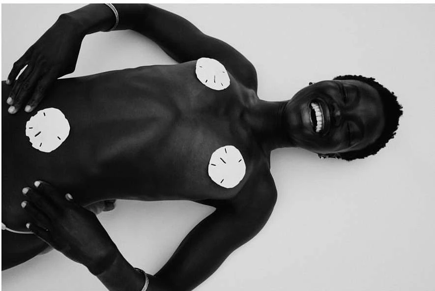 An adult woman with dark skin is lying down in a light studio. The woman is smiling with white circles on her chest and abdomen.
