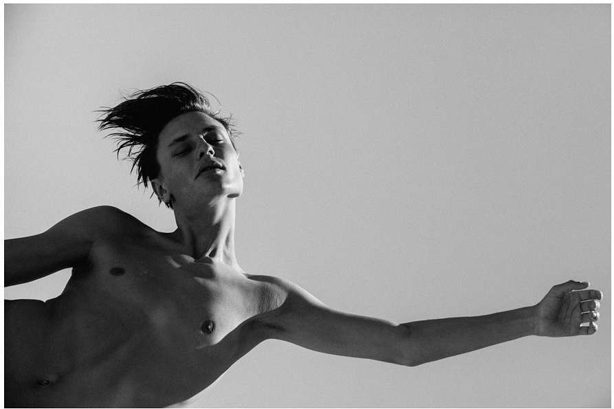 Monochromatic portrait of a bare-chested adult male in a striking pose against a stark white backdrop, artful contrast and natural light.