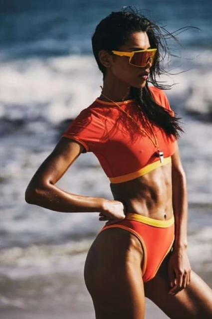 An adult female lifeguard poses in an orange two-piece swimsuit with yellow trim on a beach with ocean waves.