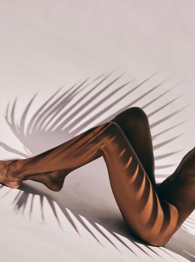 Tan-skinned legs of a woman with tropical palm leaf shadows on white surface, studio lighting, sensual, minimalist aesthetic.