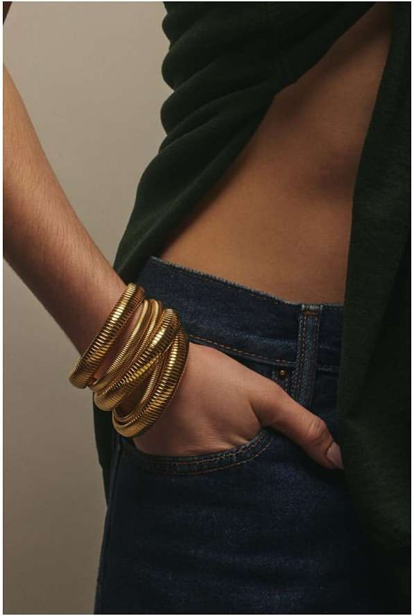 Close-up of a woman's arm wearing stacked gold bracelets, hand resting in denim jean pocket against a neutral backdrop.