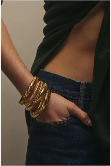 Close-up of a woman's arm wearing stacked gold bracelets, hand resting in denim jean pocket against a neutral backdrop.