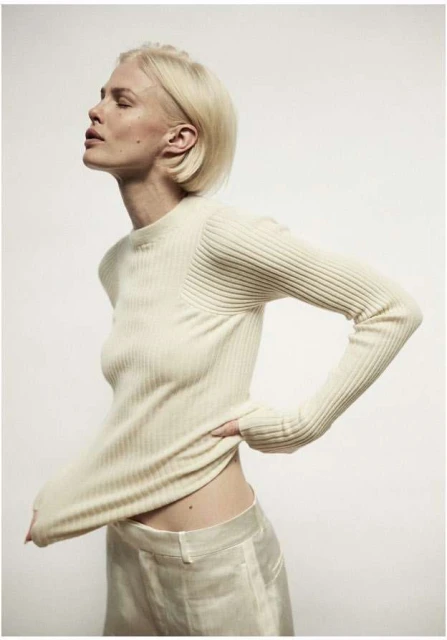 Blonde woman poses in a cream-colored ribbed sweater and matching silk pants against a bright white backdrop in a fashion shot.