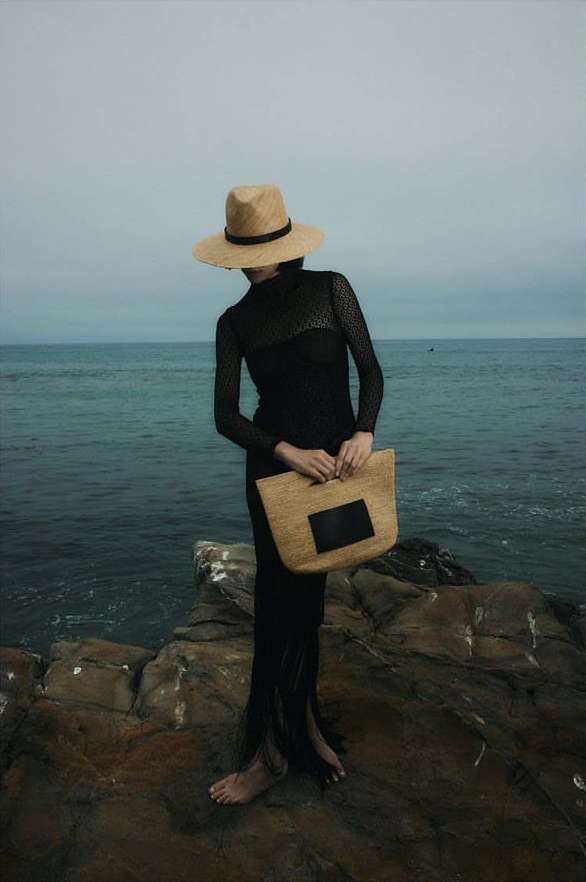 A woman in a black dress and straw hat poses on a rocky outcrop overlooking the ocean, holding a straw tote bag on an overcast day.