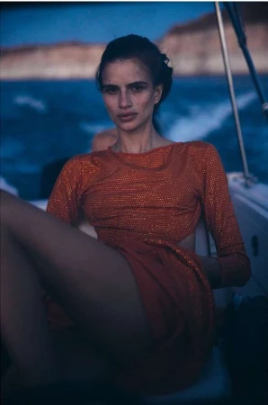 Woman in a sparkling red dress seated on a boat at dusk with water and distant coastline visible behind her.