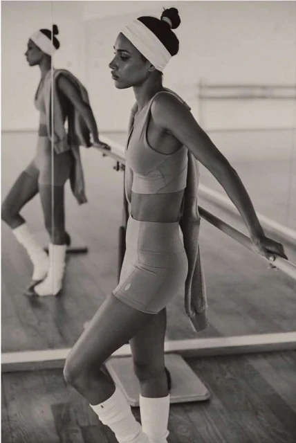 Athletic woman in sports bra and shorts stands in a mirrored dance studio with barre; warm lighting and neutral tones create a sense of focus.