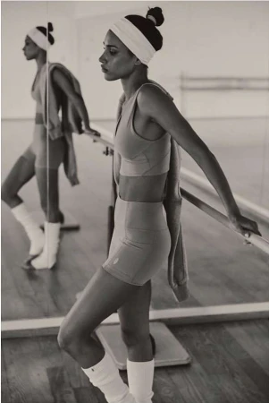 Athletic woman in sports bra and shorts stands in a mirrored dance studio with barre; warm lighting and neutral tones create a sense of focus.