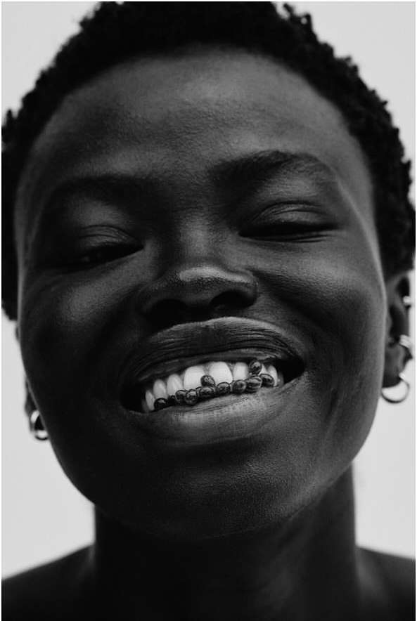 Close-up portrait of a smiling Black woman with dark short hair holding small beads in her mouth against a plain white background