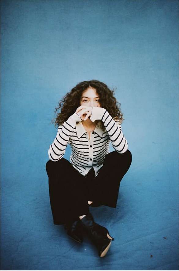 Woman with dark curly hair wearing a striped cardigan and black trousers squatting against a blue textured backdrop.