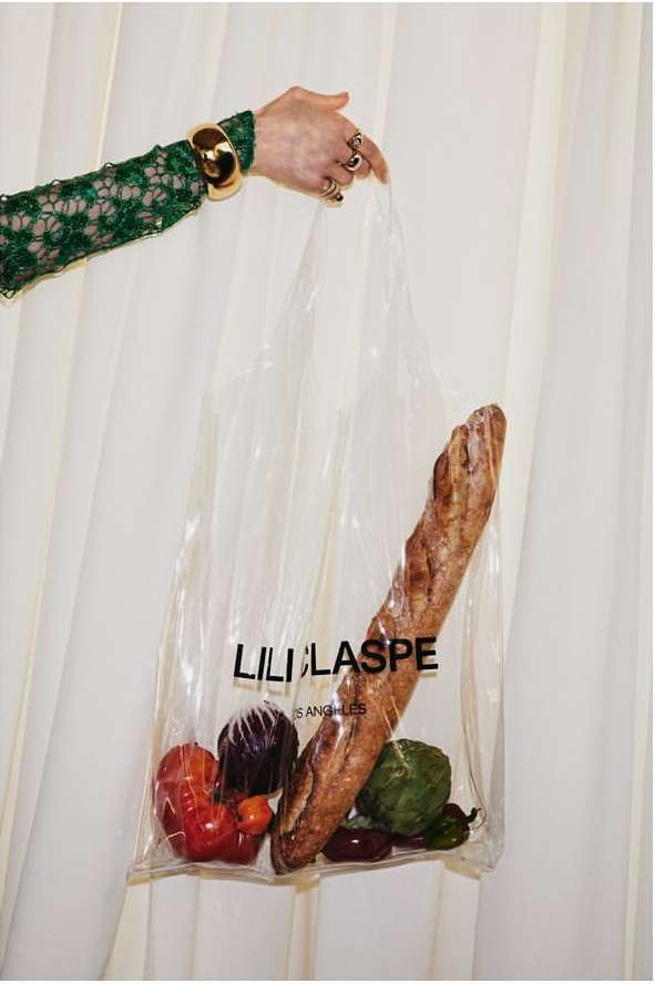 A person holds a clear plastic shopping bag filled with produce and a baguette, showcasing green sleeves with gold cuff and rings.