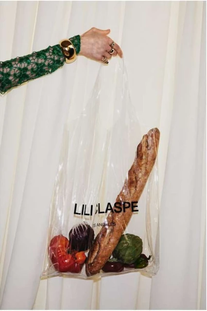 A person holds a clear plastic shopping bag filled with produce and a baguette, showcasing green sleeves with gold cuff and rings.