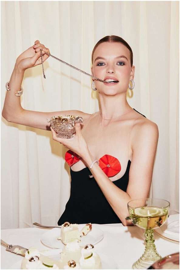 A blonde woman in a black dress holds a crystal bowl while playfully putting a silver necklace in her mouth at a table.