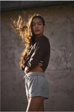 Portrait of a woman with long hair blowing in the wind wearing a dark top and grey shorts against a concrete wall