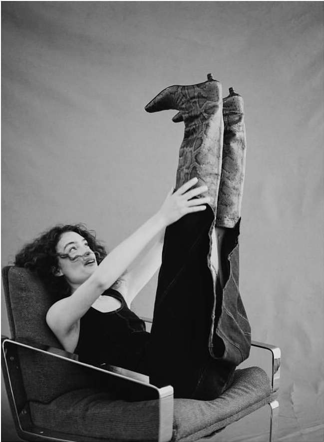 Woman seated in an armchair lifting her legs up in tall patterned boots, photographed in black and white by Aaron Feaver.