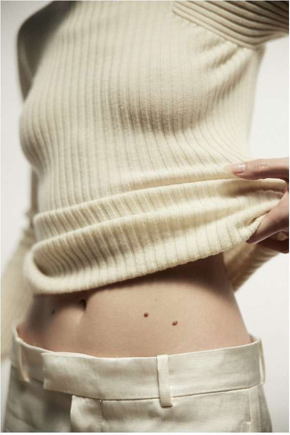 Close-up shot of a light-skinned adult female wearing a ribbed knit sweater and white pants in an indoor setting against a white backdrop.