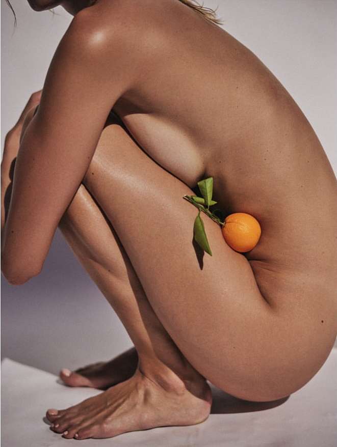 Woman in a squatting position with an orange against her hip. The background is white. The light source is on the top.