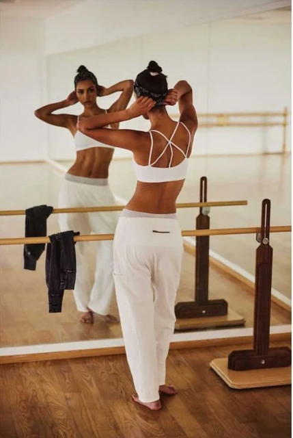 Athletic woman in white activewear poses in front of a mirror in a dance studio, showcasing her back and toned physique in fitness apparel.