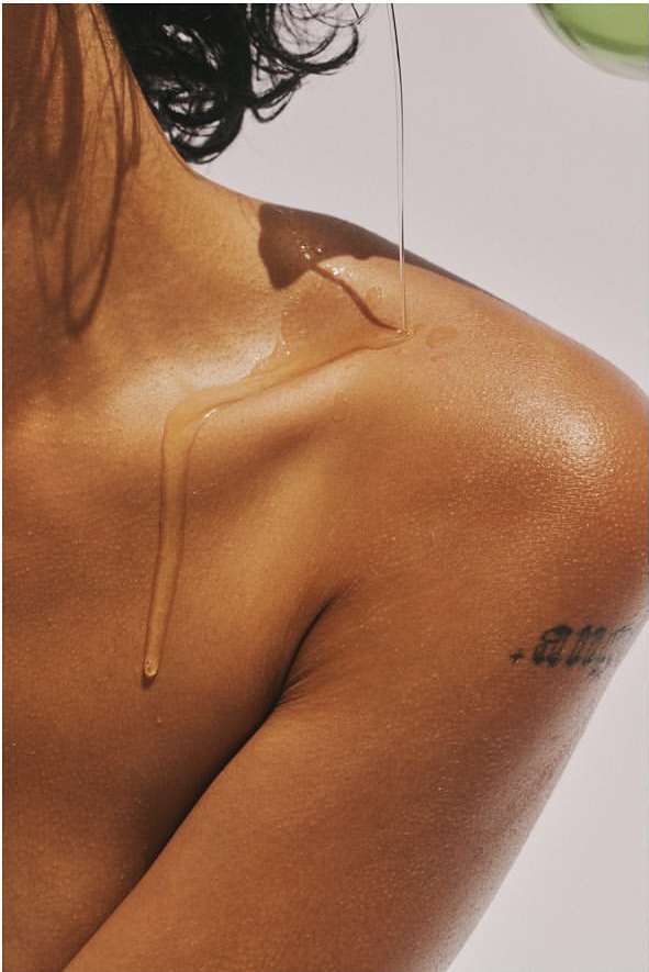 Close-up of a person's shoulder and upper arm as oil is poured over their skin, highlighting texture and a tattoo against a light background.