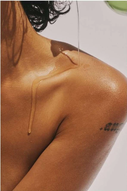 Close-up of a person's shoulder and upper arm as oil is poured over their skin, highlighting texture and a tattoo against a light background.