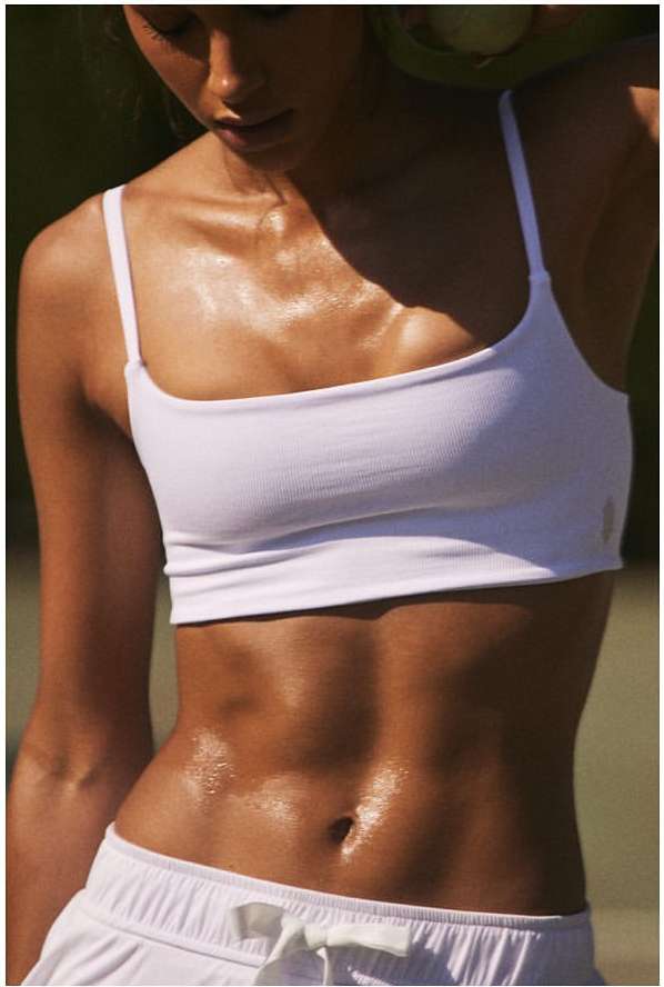 Adult woman with muscular physique wearing a white sports bra and shorts, standing in a sunny outdoor setting with a green backdrop.