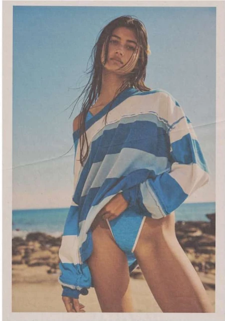 Woman in blue bikini and striped oversized top poses on a beach with blue sky and ocean in background. Fashion, swimwear.