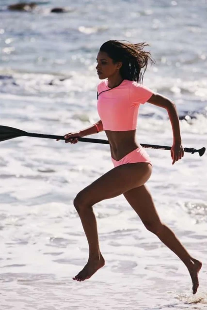 Athletic woman in pink swimwear running barefoot on a beach with a paddle in hand, ocean waves, and bright daylight.