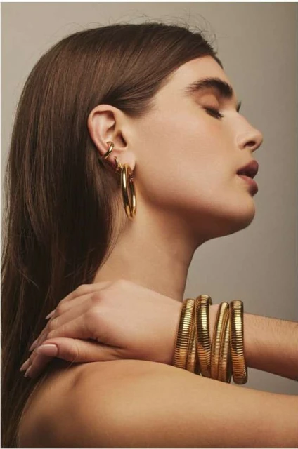 Woman with closed eyes poses in profile wearing large gold hoop earrings and multiple stacked coil bracelets in a studio environment.