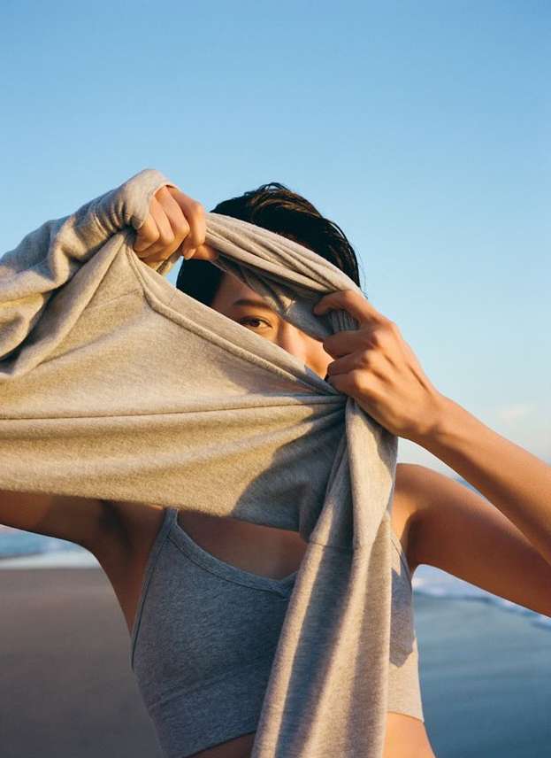 Woman holding up grey sweatshirt on the beach with blue sky and water in the background; fitness and outdoor lifestyle concept.