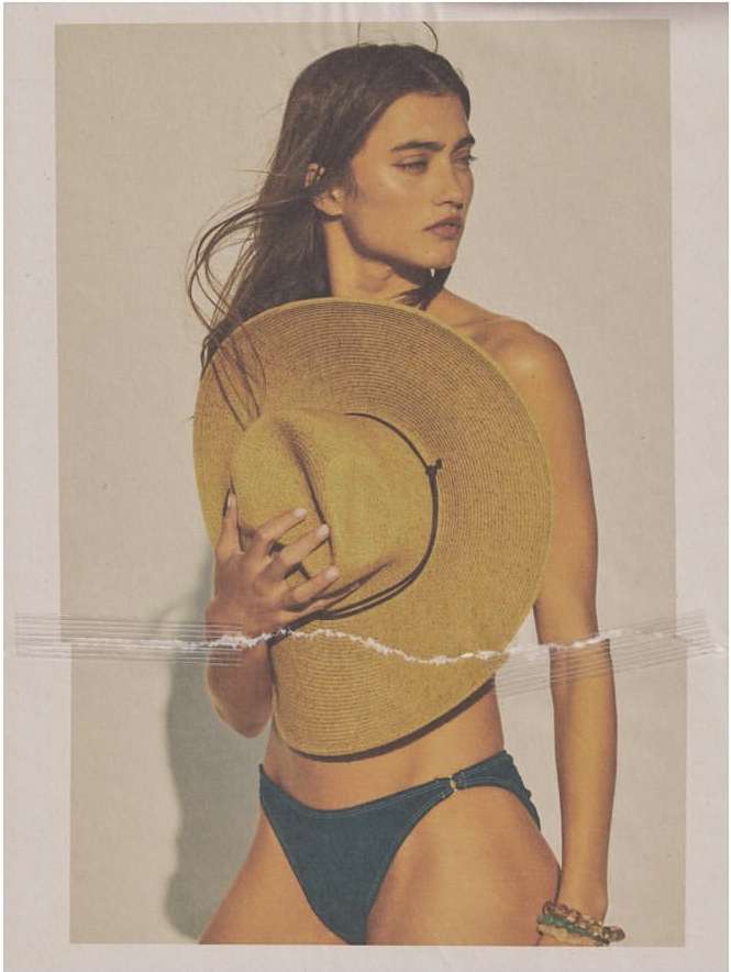 An adult woman poses in a studio, wearing a bikini bottom while holding a straw hat in front of her torso against a white background.