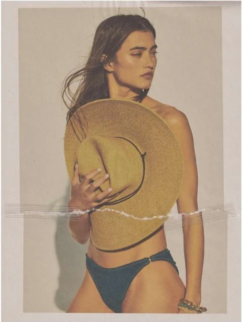 An adult woman poses in a studio, wearing a bikini bottom while holding a straw hat in front of her torso against a white background.