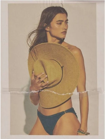 An adult woman poses in a studio, wearing a bikini bottom while holding a straw hat in front of her torso against a white background.