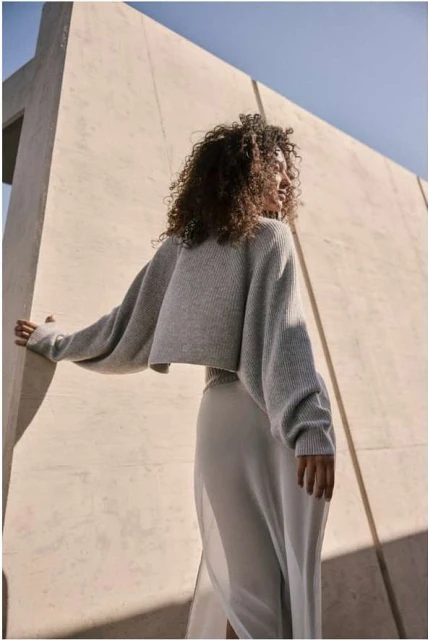 An adult female with curly hair poses in a gray sweater and light-colored skirt, against a geometric architectural backdrop under a sunny sky.