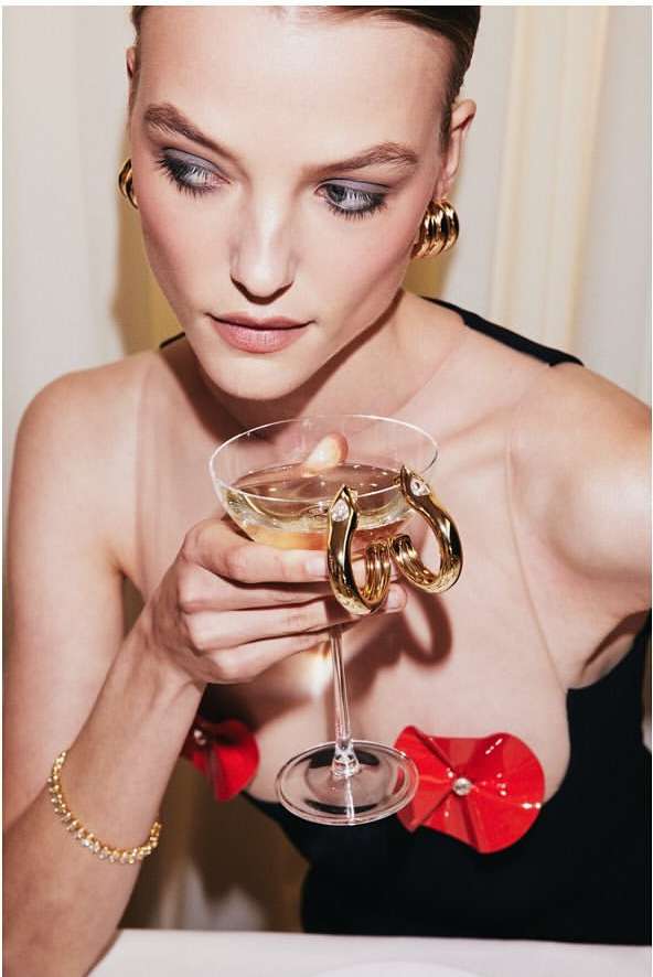 Elegant woman in a black dress with red flower accents holds a champagne coupe, showcasing gold jewelry in a brightly lit setting.