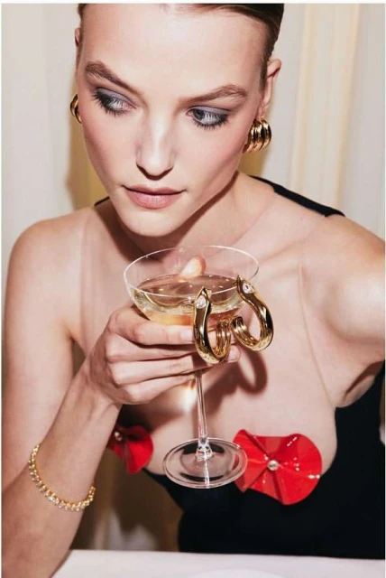 Elegant woman in a black dress with red flower accents holds a champagne coupe, showcasing gold jewelry in a brightly lit setting.