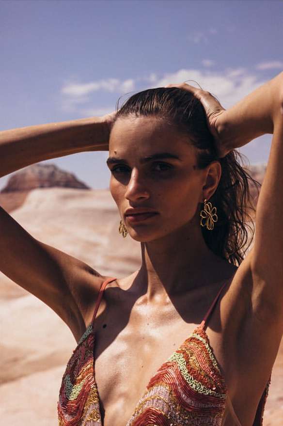 Woman poses in the desert wearing flower earrings and a sequined top, her hands are up behind her head, rocks and blue sky visible.