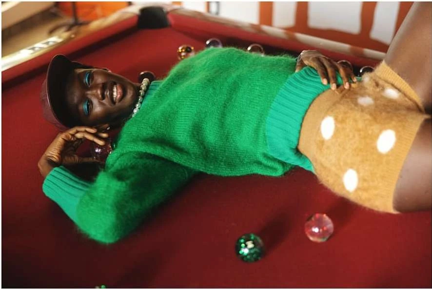 Woman in green sweater and cap poses on red pool table with billiard balls, creating a vibrant and playful fashion-forward image.