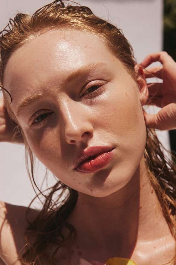 Close-up portrait of a model with wet red hair and fair skin posing with her hands behind her head in a brightly lit photo studio.