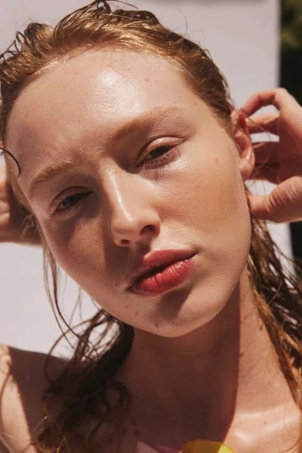Close-up portrait of a model with wet red hair and fair skin posing with her hands behind her head in a brightly lit photo studio.