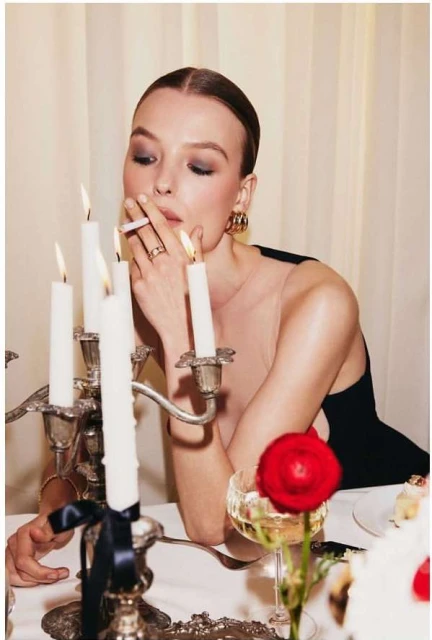 Elegant woman smoking at dinner table with candelabra, wine glass, flower, and plate with dessert in soft indoor lighting.