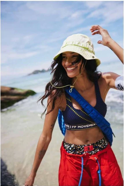 Smiling woman in a bucket hat and workout gear on a sunny beach. She wears a navy sports bra, red pants, and a blue backpack.