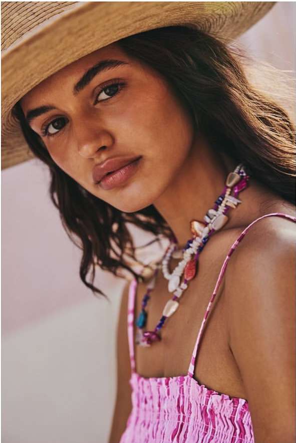 Close-up of a woman with dark hair wearing a straw hat and pink top, adorned with colorful beaded necklaces, against a soft pink background.