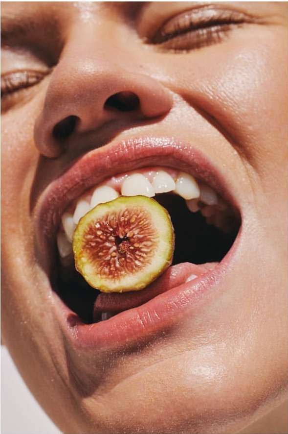 Close-up of a woman holding a fig in her mouth, bright sunny lighting, light skin tone, smooth skin texture, bright white teeth.