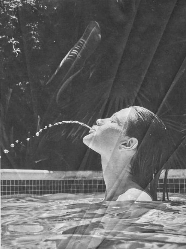 Woman in a swimming pool drinking water shot into the air, captured in black and white with a tropical background of trees and plants.
