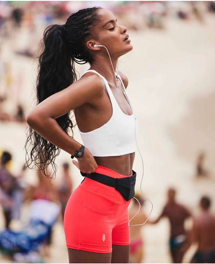 Black woman wearing a white sports bra and coral shorts on a sandy beach, photographed by Aaron Feaver.