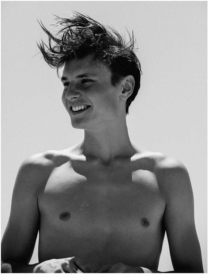 Monochrome portrait of a young adult man with tousled hair, smiling shirtless under bright daylight. Head and shoulders shot. 
